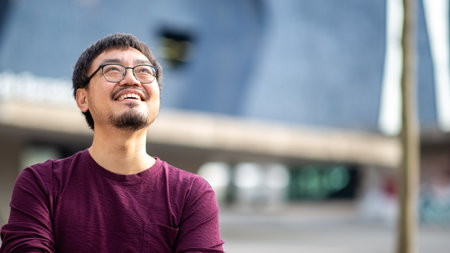 An Asian man in glasses and a maroon shirt looking upwards with a smile, standing outdoors in an urban area with blurred modern architecture in the backgroundの写真素材