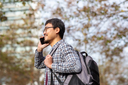 Side view of an Asiatic man in glasses and a plaid shirt talking on a phone while carrying a...