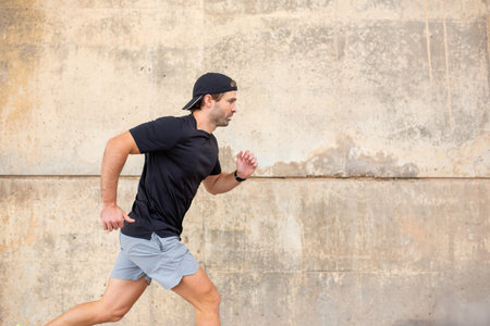 Male athlete in black t-shirt and gray shorts running along a concrete wall outdoors, showcasing...