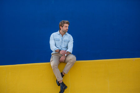 A man in a light denim shirt, beige pants, and black boots sits on a yellow wall with a solid blue backgroundの写真素材