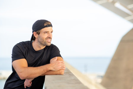 Man in sportswear resting against a concrete wall, smiling and looking into the distance, with the...