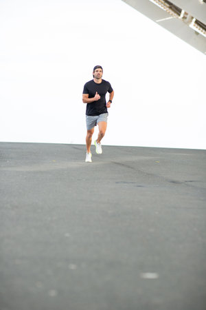 A man in a black t-shirt, gray shorts, and white sneakers running on a paved surface as part of his...