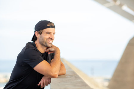 Portrait of a cheerful man in sportswear leaning on a concrete wall and smiling, with the ocean and...