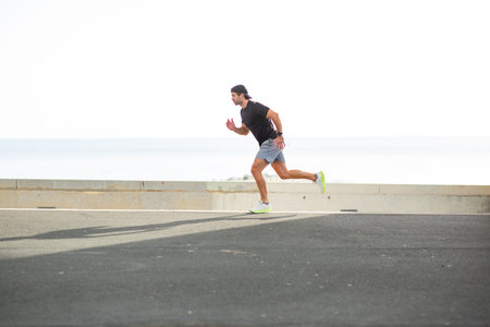 Full-body side view of a man running along a paved road with the ocean and rocks in the background...