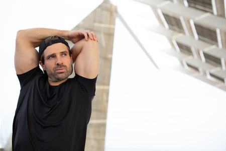 Close-up of a man in a black t-shirt and cap stretching his arms overhead outdoors, surrounded by...