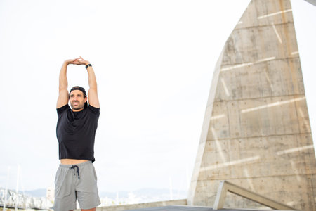 A man wearing a black t-shirt and gray shorts, stretching his arms overhead outdoors with modern...