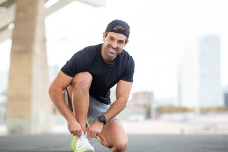A man in a black t-shirt, gray shorts, and white sneakers crouching and smiling as he ties his shoes...