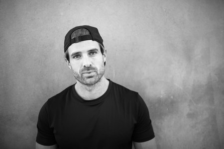 Monochrome close-up of a man wearing a black t-shirt and backward cap, standing against a plain wall...