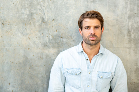 A close-up color portrait of a man wearing a light denim shirt standing outdoors in front of a...