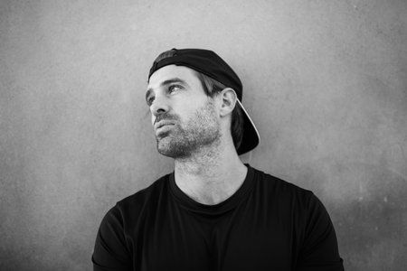 Monochrome close-up of a man wearing a black t-shirt and backward cap looking up and to the side...