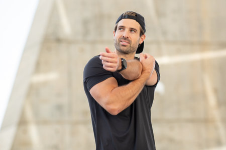 Close-up of a man wearing a black t-shirt and cap performing an arm stretch outdoors with modern...
