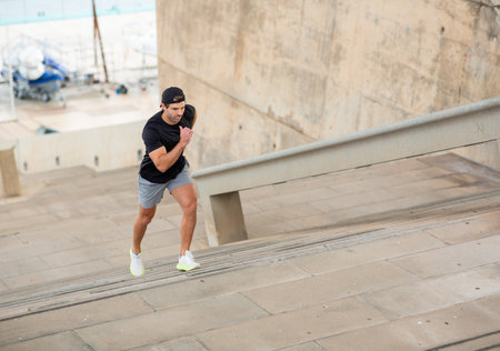 A man in a black t-shirt, gray shorts, and white sneakers running up a set of stairs outdoors with a...