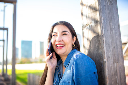 A cheerful woman wearing a denim shirt is talking on her smartphone while leaning against a wooden...