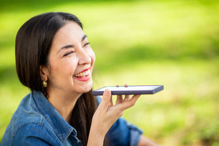 A woman wearing a denim shirt is outdoors in a park, holding her smartphone close to her mouth while...