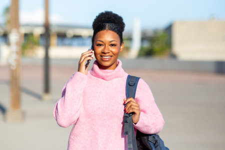 A young woman wearing a pink sweater and carrying a backpack smiles while talking on her smartphone...