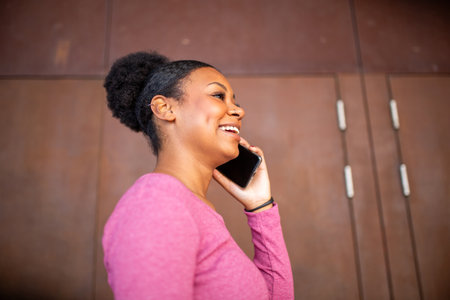 A young woman in a pink t-shirt smiling and talking on a smartphone while standing near a brown...