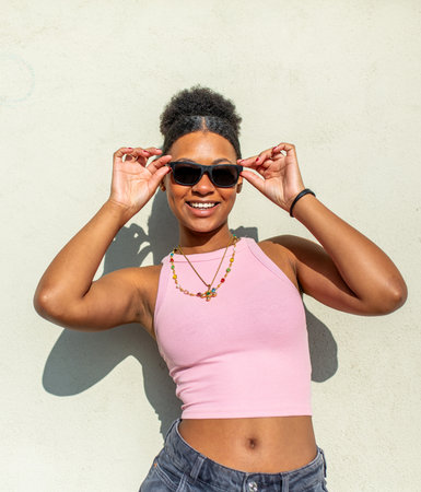 Woman wearing a pink crop top and layered necklace, smiling and adjusting sunglasses with both hands...