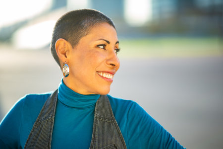 A confident woman with short hair and hoop earrings wearing a blue turtleneck and a striped vest smiles warmly while looking to the side outdoorsの写真素材