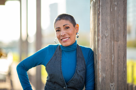 Woman wearing a blue turtleneck and pinstripe vest smiling while leaning on a wooden post in a sunny...