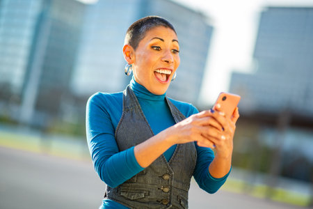 A cheerful woman in a blue turtleneck and gray vest looking at her smartphone with a joyful...