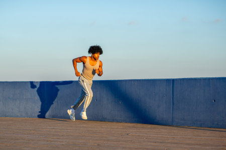 Athletic young man in a tank top and sweatpants runs with intensity along a wooden boardwalk next to a blue wall during golden hourの写真素材