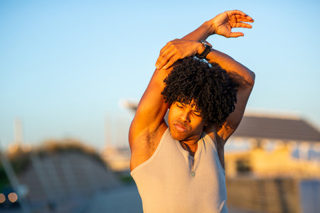 Close-up of a young man with curly hair stretching his arm overhead while bathed in warm sunlight,...