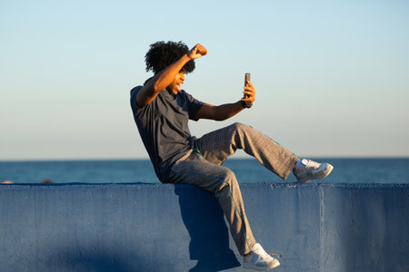 Young man with curly hair sits on a blue wall by the ocean, raising his arm with excitement while...