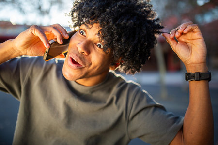 A young man animatedly talks on the phone outdoors, holding his hair and showing a surprised expression while enjoying a lively conversation