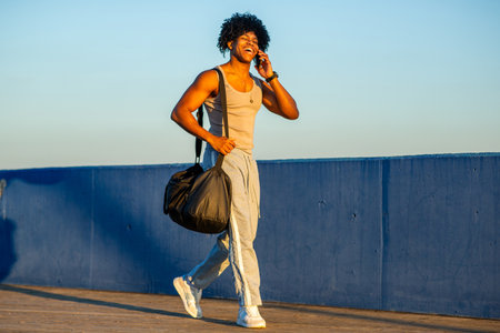 Confident young man in activewear talking on the phone with a big smile while walking along a blue seaside promenade. He carries a black gym bag, enjoying a sunny day by the waterの写真素材