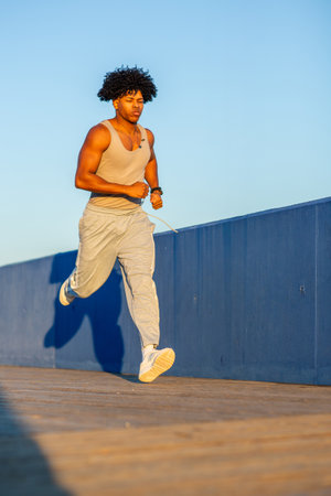 Fit young man in a tank top and sweatpants running on a boardwalk during golden hour. Focused...