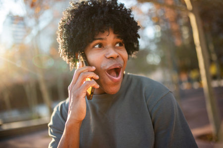 Young man with curly hair shows a surprised expression while speaking on his smartphone outdoors in warm sunlightの写真素材
