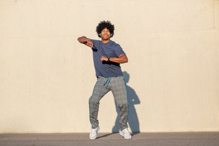 Smiling young man with headphones dances playfully in the street, enjoying music in a casual outfit...