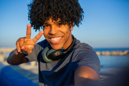 Happy young man at the beach flashes a peace sign while taking a selfie, wearing headphones around...