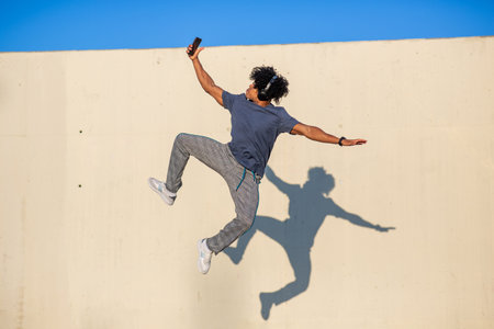 Man wearing headphones jumps into the air holding a smartphone, captured mid-leap against a sunlit...