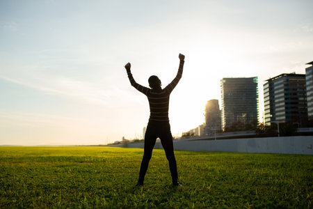A silhouette of a woman with short hair wearing a striped sweater, standing in a park with her arms...