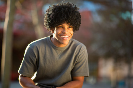 Young man with curly hair smiles warmly while leaning forward outdoors, bathed in soft sunlight and...