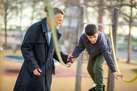 Senior man in a coat carefully helping a child walk across a rope structure at a playground on a cool dayの写真素材