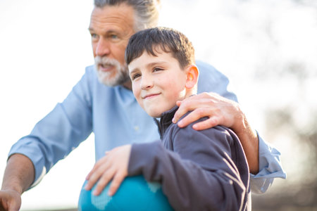 A happy boy holding a blue ball smiles while standing next to his proud grandfather in a bright and cheerful park settingの写真素材