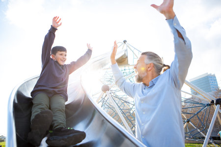 Boy throws his hands up in excitement while sliding down a playground slide to meet his joyful grandfatherâs high-five on a sunny dayの写真素材