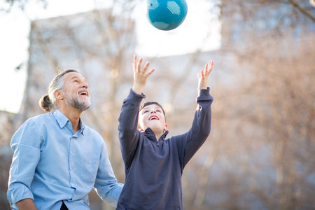 Grandson throws a blue ball into the air while grandfather looks on with joy, enjoying a playful moment in a sunny outdoor parkの写真素材