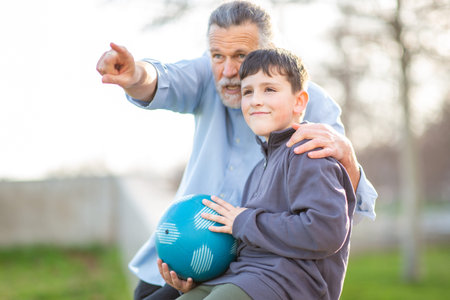 Grandfather points into the distance while the young boy holds a blue soccer ball, both smiling and enjoying a calm outdoor momentの写真素材