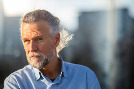 Portrait of an older man with a serious expression and direct gaze, standing outdoors in sunlight wearing a blue shirt with a modern urban backgroundの写真素材