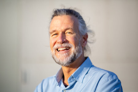 Elderly man smiling brightly in natural light, wearing a light blue shirt and standing outdoors with a plain background and relaxed demeanorの写真素材
