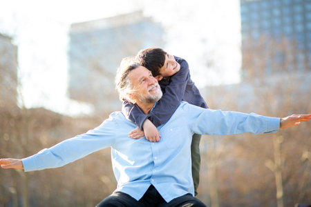 An older man spreads his arms wide like airplane wings while his smiling grandson hugs him from behind, both enjoying a fun moment in the parkの写真素材