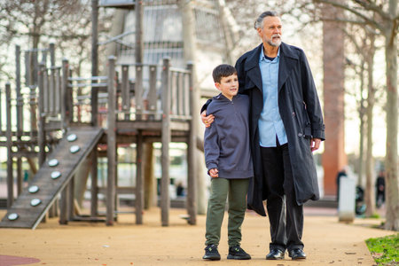 Elderly man walking with a young boy, arm around his shoulders, near a large wooden playground structure in a parkの写真素材