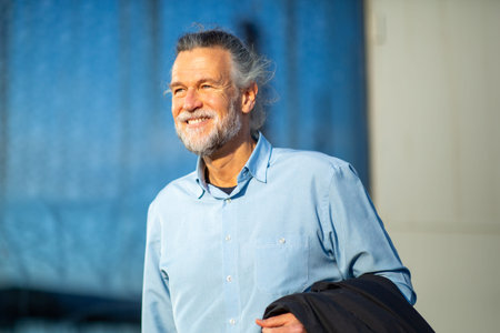 Happy senior man in a light blue shirt smiling while holding a jacket over his shoulder on a sunny dayの写真素材