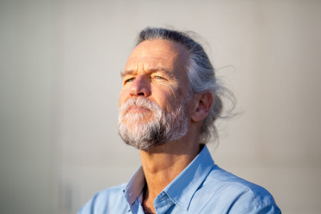 Close-up of a serious senior man with a beard looking upward with a thoughtful expression under natural sunlightの写真素材