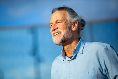 Cheerful elderly man with gray hair and beard smiling joyfully under a clear blue sky, wearing a light blue shirt on a sunny dayの写真素材