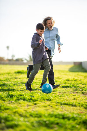 Young boy runs joyfully with his grandfather chasing a blue soccer ball across a sunny park fieldの写真素材