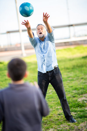 Energetic grandfather tosses a blue ball with both hands toward his grandson while playing together on a sunny green fieldの写真素材